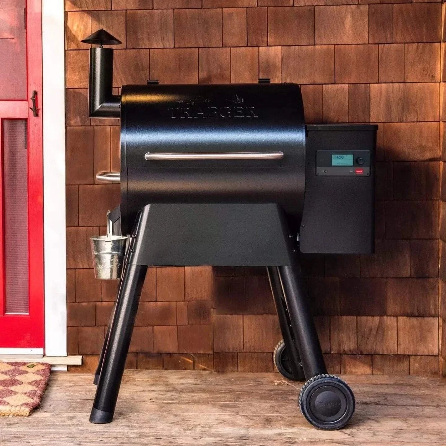Black Traeger grill against a wooden wall with a red door.