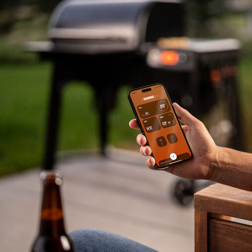 Person using a smartphone app to control a grill with a beer bottle on a table in the foreground.