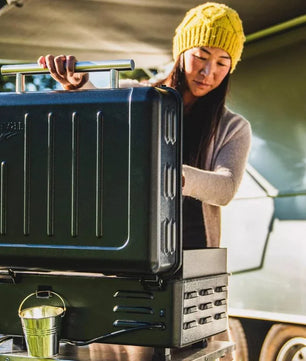 Person opening a black camping stove in front of a camper van