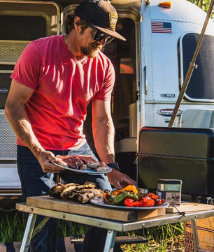 Man grilling outdoors next to a trailer with food on a table
