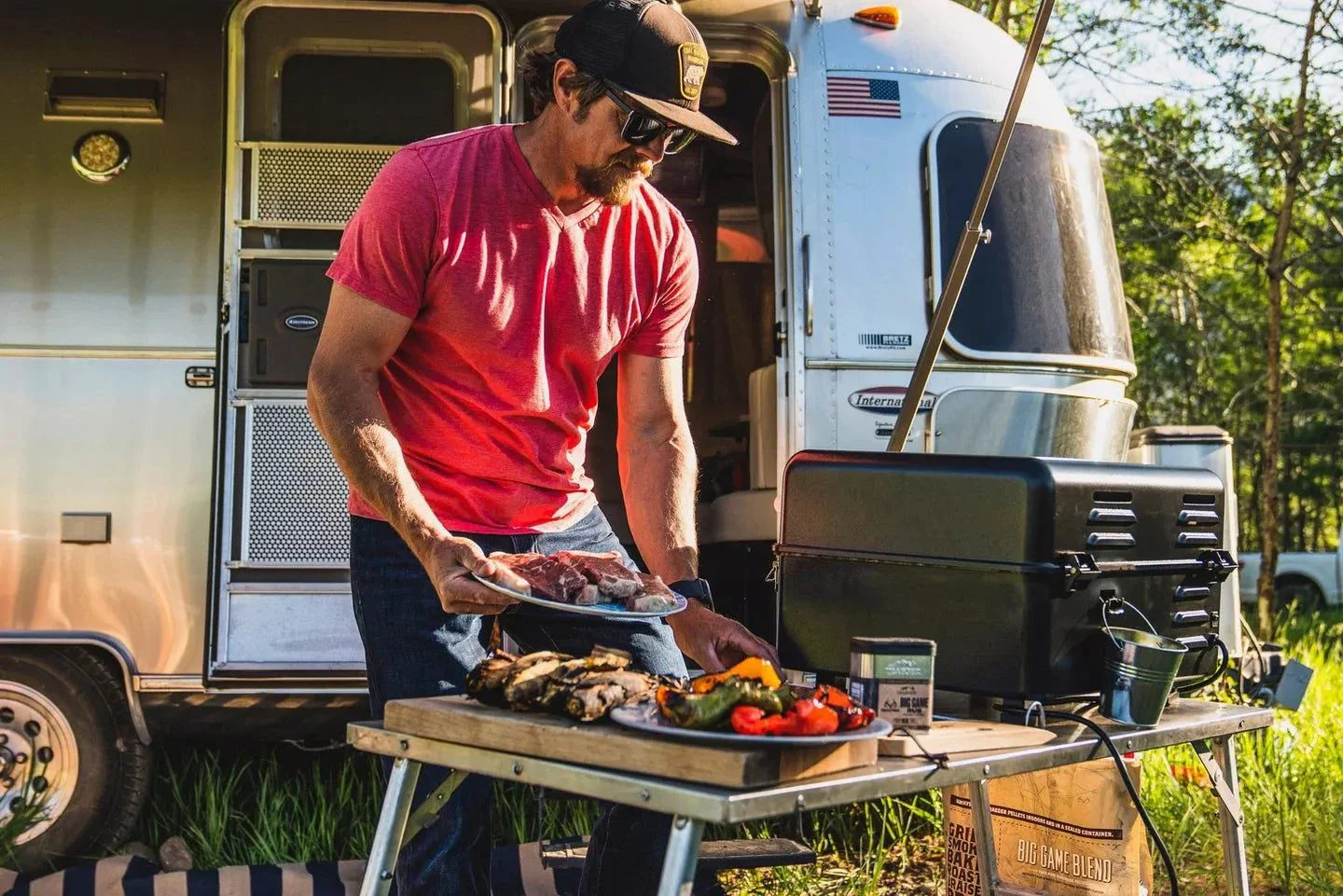 Man grilling outdoors next to a trailer with food on a table