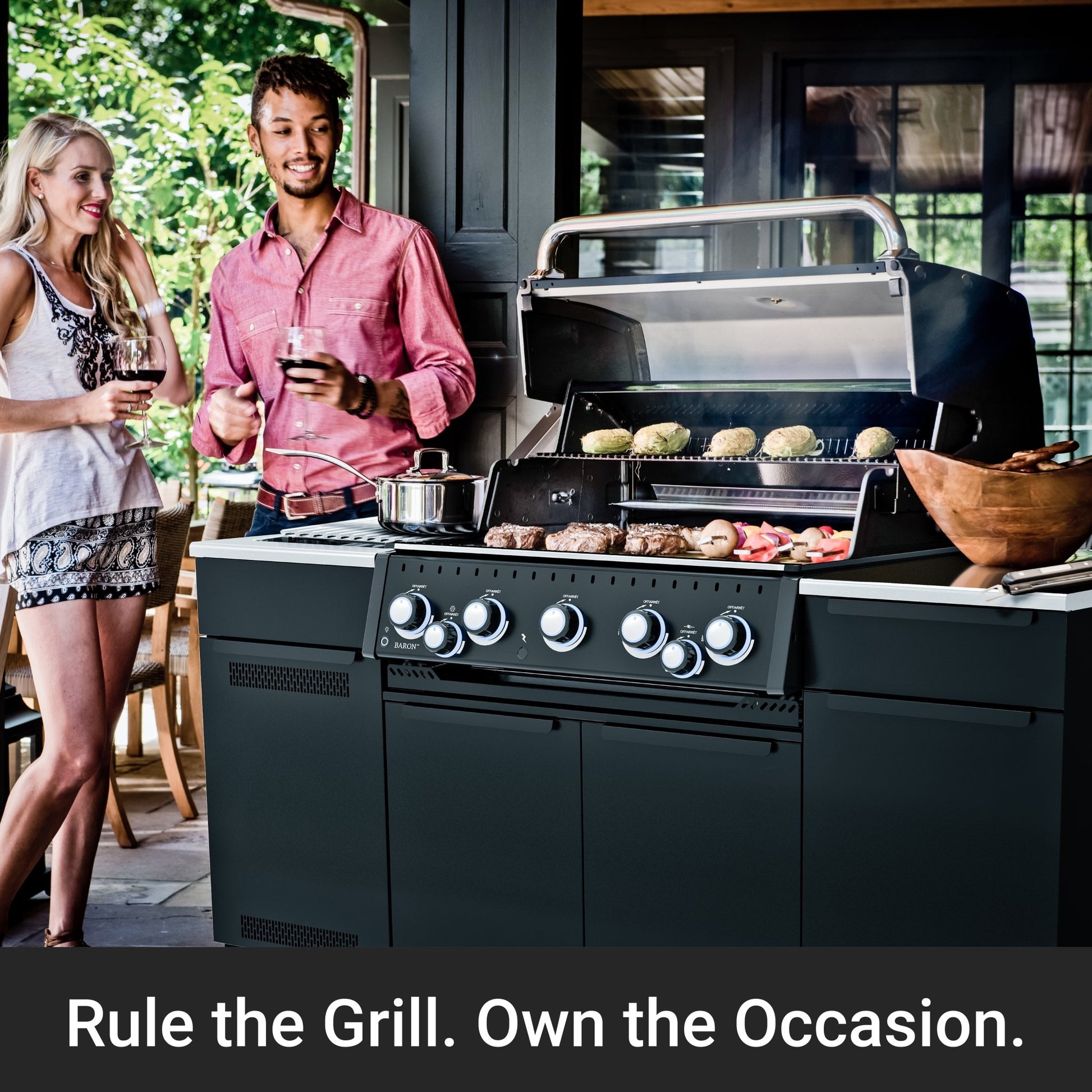 A large black outdoor gas grill with six control knobs is shown open, cooking meat and burgers on a patio. Two people holding wine glasses stand to the left, and a dark promotional banner at the bottom reads, 'Rule the Grill.