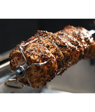 A close-up of a seasoned meat skewer rotating on a metal spit roast fork, with a dark, blurred background.