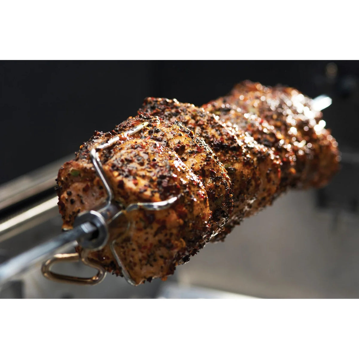 A close-up of a seasoned meat skewer rotating on a metal spit roast fork, with a dark, blurred background.