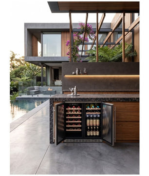 A built-in undercounter wine refrigerator with glass doors, shown open with wine bottles inside, set in a modern outdoor kitchen/bar area next to a pool with a contemporary house in the background.