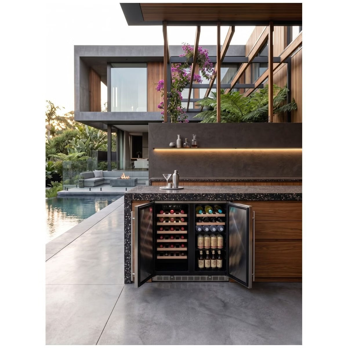 A built-in undercounter wine refrigerator with glass doors, shown open with wine bottles inside, set in a modern outdoor kitchen/bar area next to a pool with a contemporary house in the background.