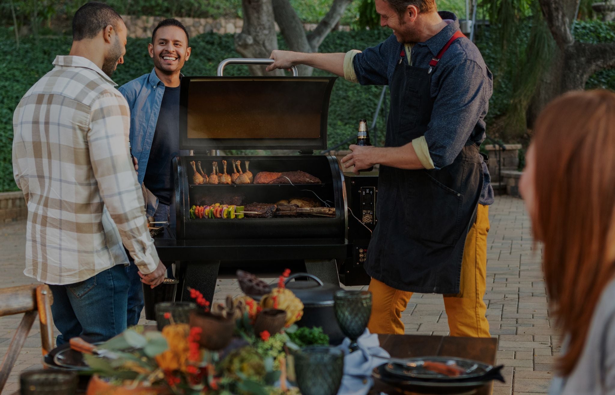 A group of people gathered outdoors around a black charcoal grill, with one person opening the lid and another holding a bottle. A dining table with food and drinks is visible in the foreground, and trees and patio paving are in the background.