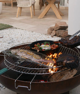 Outdoor grill with food being cooked on a patio with chairs and a table in the background.
