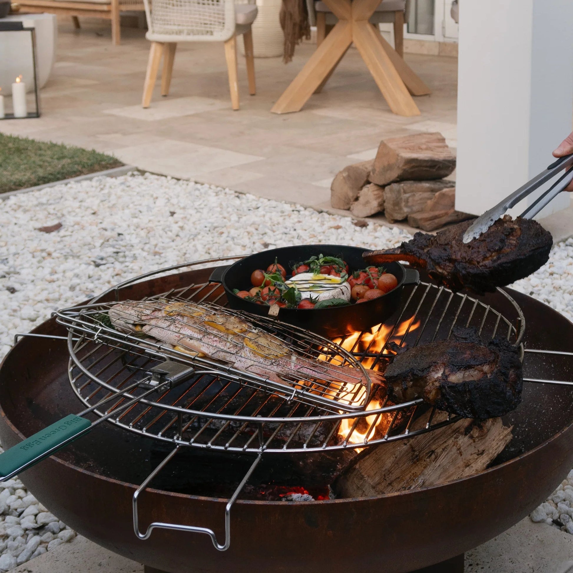 Outdoor grill with food being cooked on a patio with chairs and a table in the background.