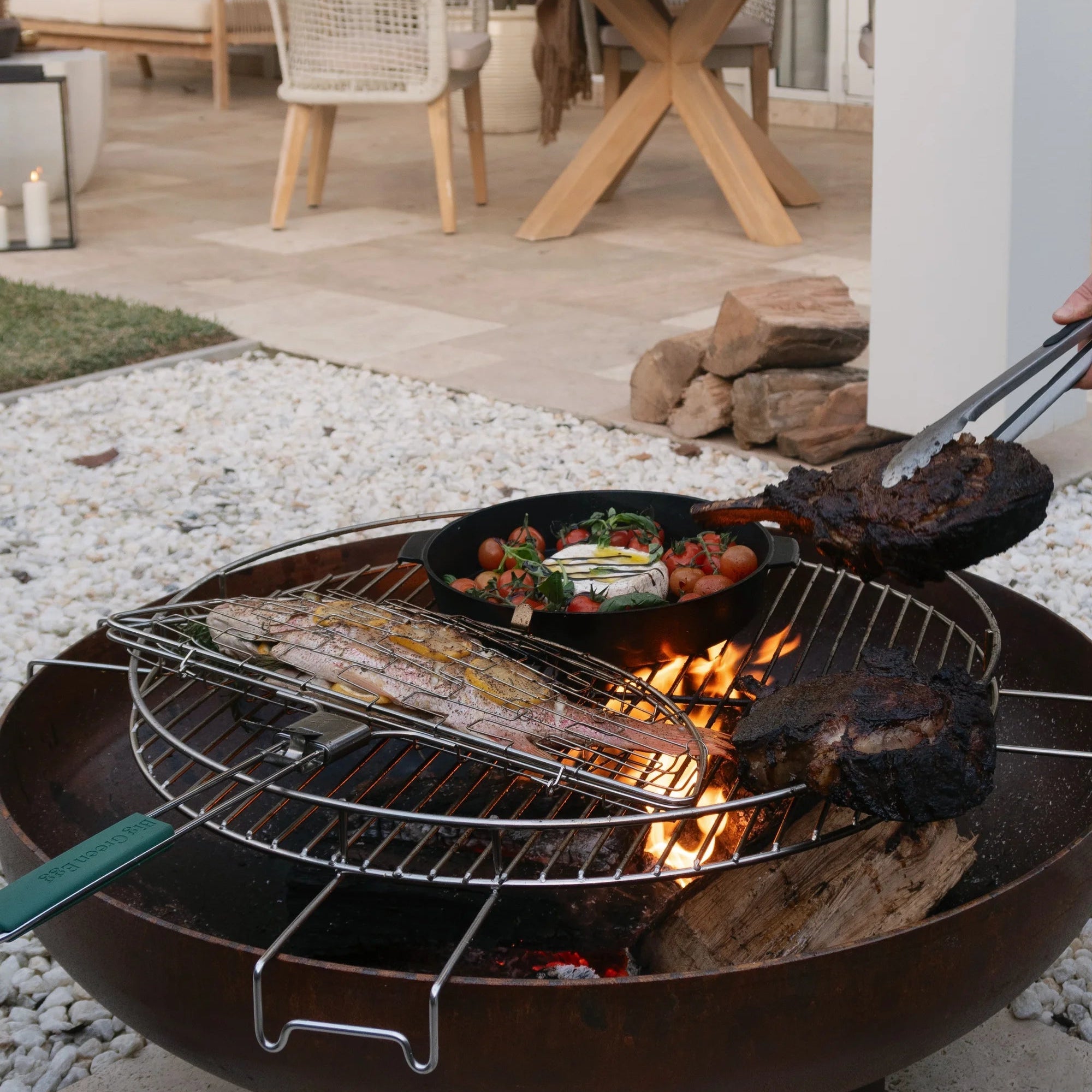 Outdoor grill with food being cooked on a patio with chairs and a table in the background.