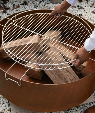 Person setting up a fire pit with a grill grate on a pebbled surface.