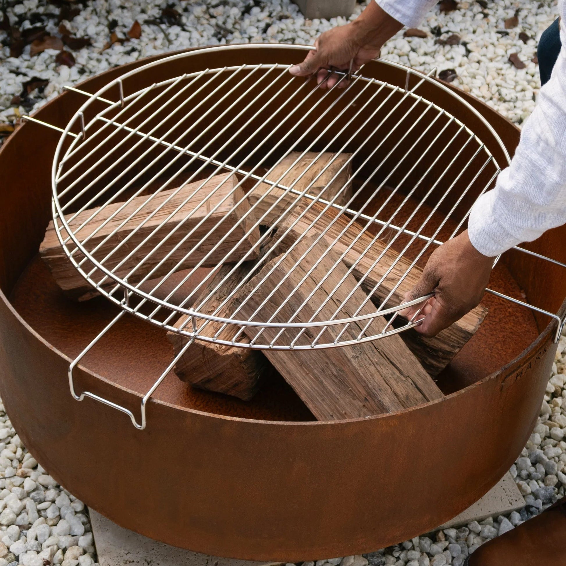 Person setting up a fire pit with a grill grate on a pebbled surface.