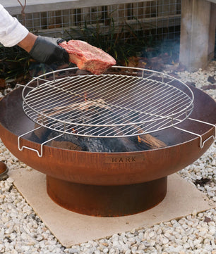 Person grilling a steak on a Hark fire pit grill outdoors.