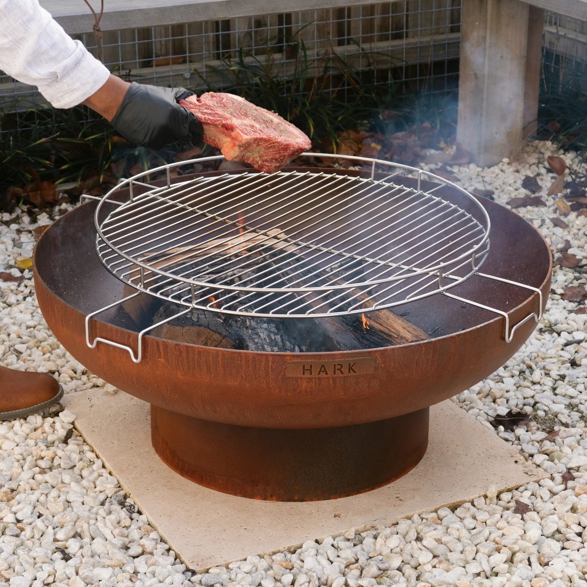 Person grilling a steak on a Hark fire pit grill outdoors.