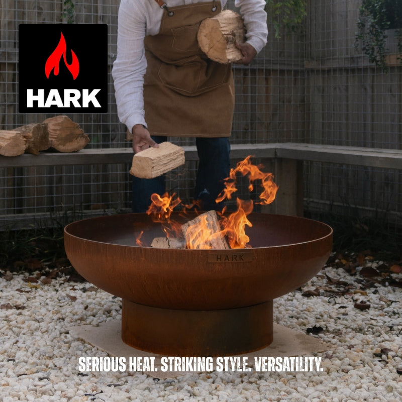 A person in a brown apron and white shirt places logs into a large round rusted metal fire pit bowl outdoors on white gravel, with visible flames. The HARK logo appears in the top left, and the text 'SERIOUS HEAT.