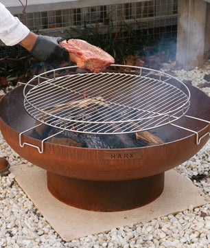 Person grilling a steak on a Hark fire pit grill outdoors.