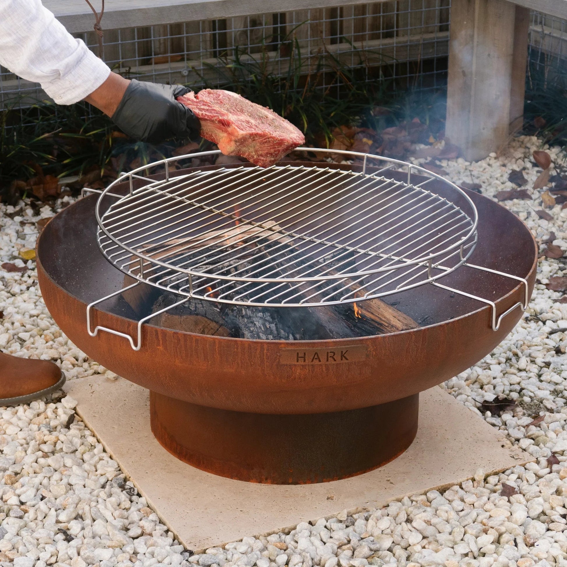 Person grilling a steak on a Hark fire pit grill outdoors.