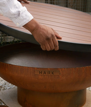 Person lifting a wooden lid off a fire pit with 'Hark' branding.