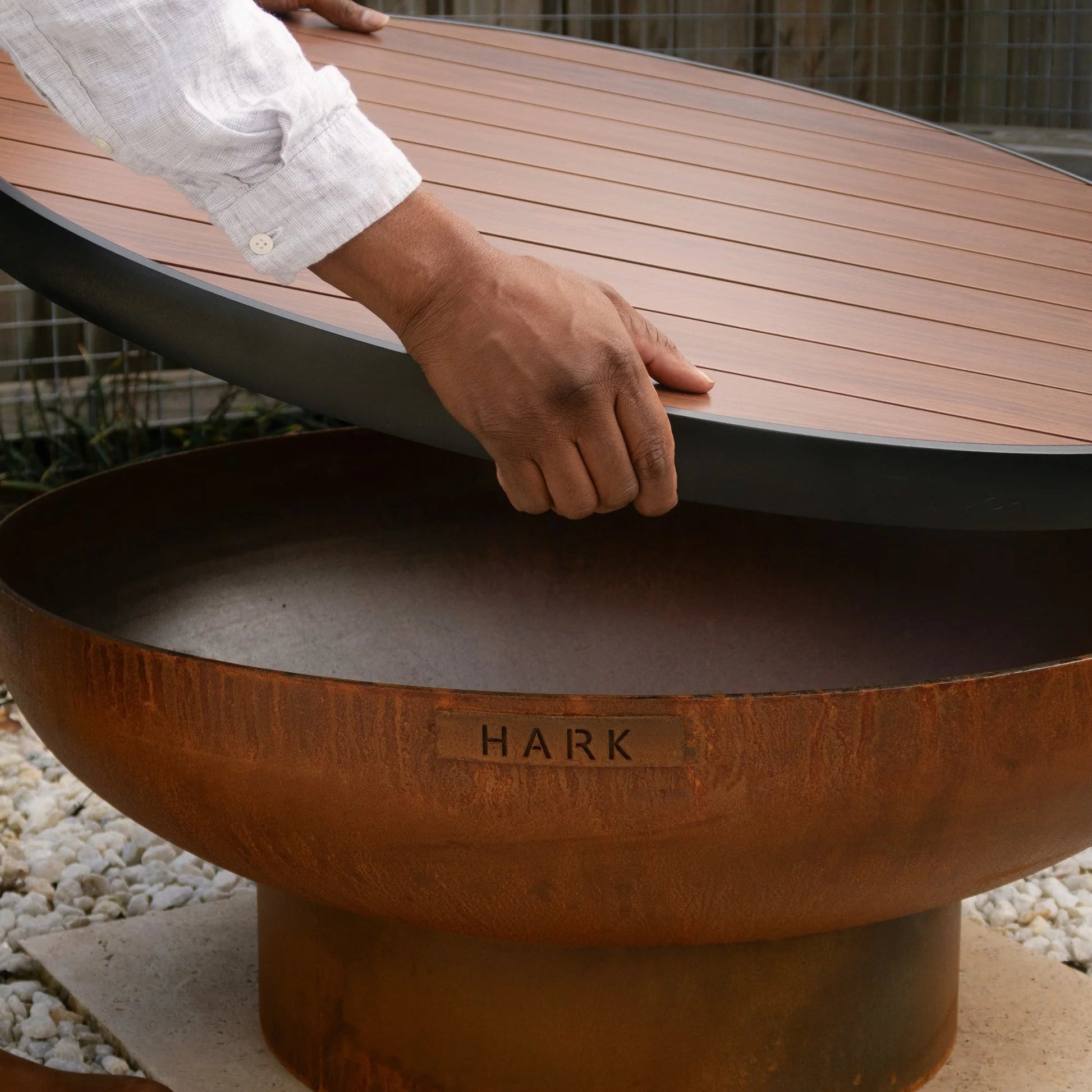 Person lifting a wooden lid off a fire pit with 'Hark' branding.