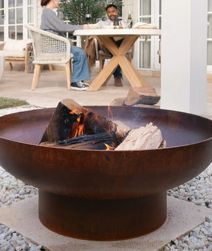 Rust-colored fire pit with flames on a pebble-covered patio, people sitting at a table in the background.