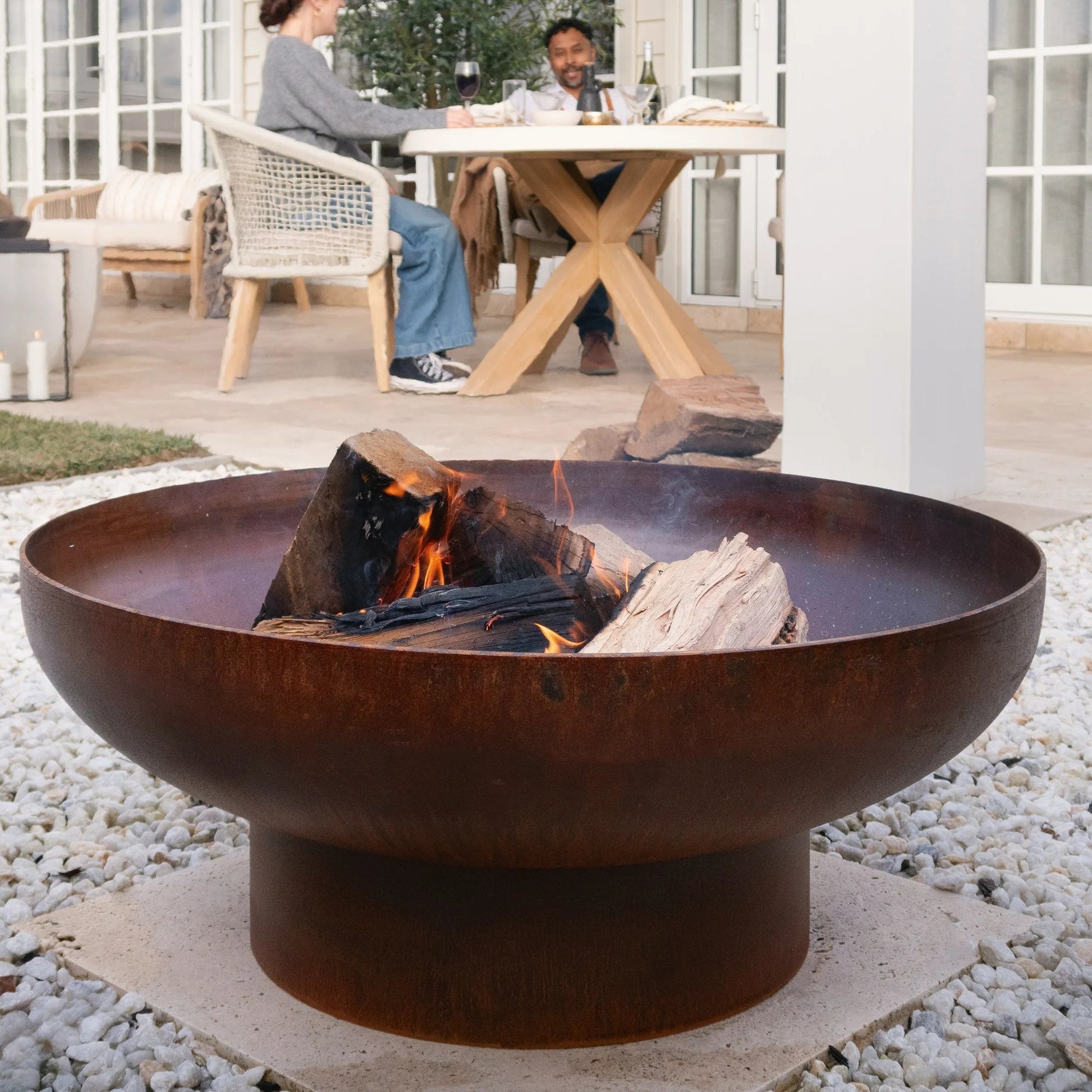 Rust-colored fire pit with flames on a pebble-covered patio, people sitting at a table in the background.