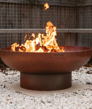 Rust-colored fire pit with flames burning on a gravel surface.