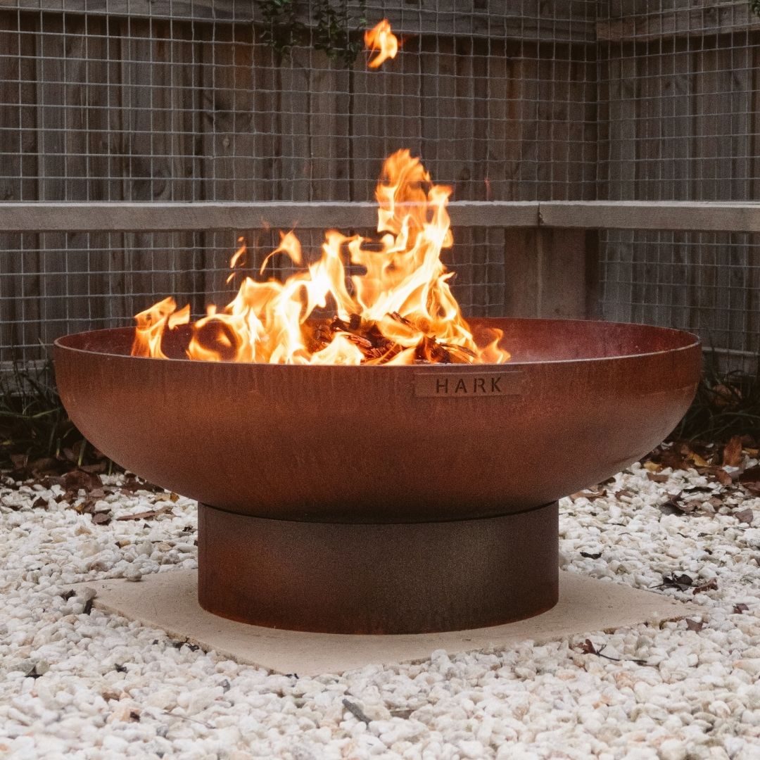 Rust-colored fire pit with flames burning on a gravel surface.