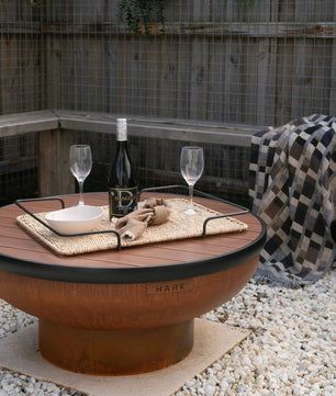 Outdoor setting with a fire pit table, wine glasses, and a bottle on a gravel surface.