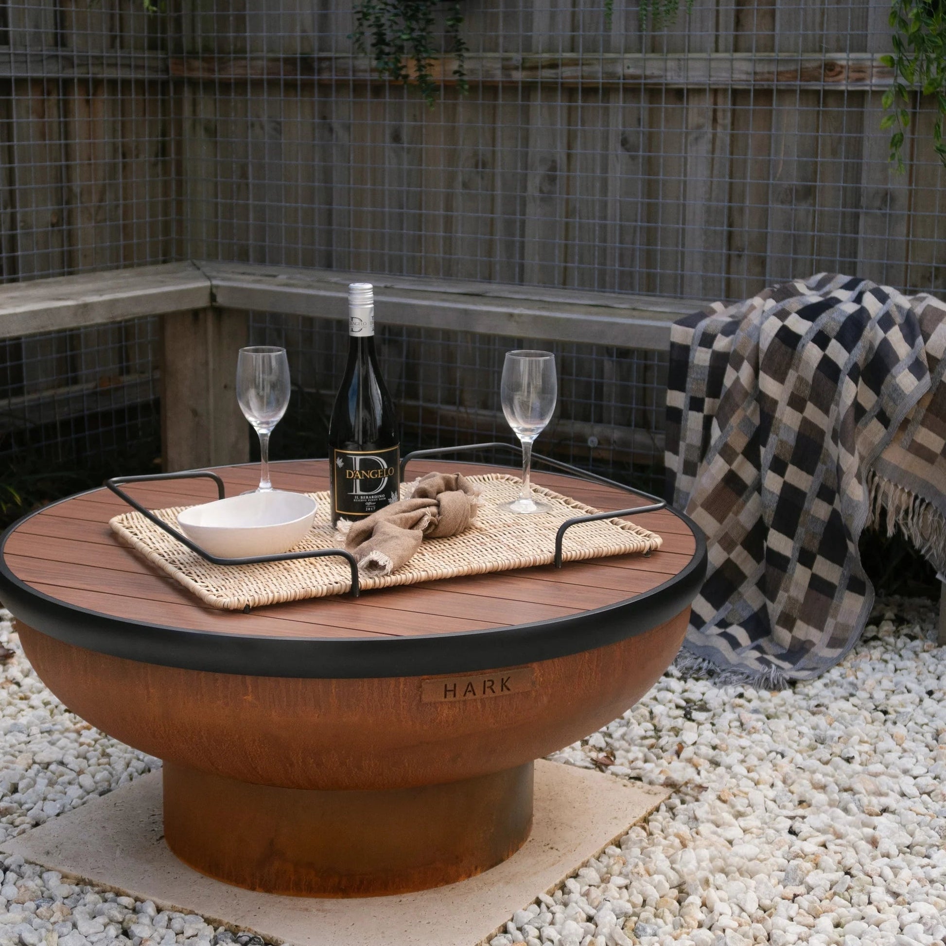 Outdoor setting with a fire pit table, wine glasses, and a bottle on a gravel surface.