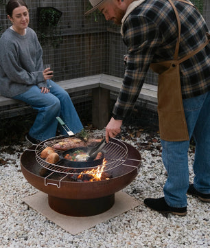 Man grilling food on a fire pit while a woman watches in an outdoor setting.