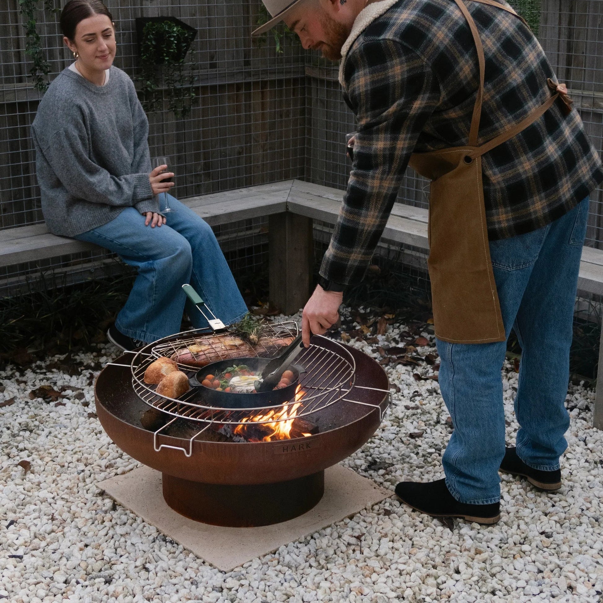 Man grilling food on a fire pit while a woman watches in an outdoor setting.