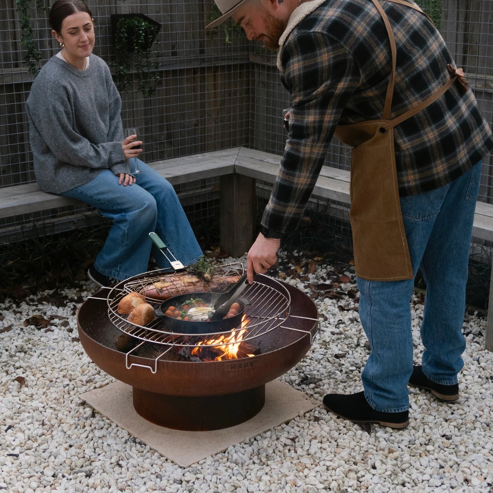 Man grilling food on a fire pit while a woman watches in an outdoor setting.