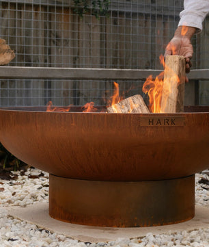 Person lighting wood in a rust-colored fire pit with a garden background