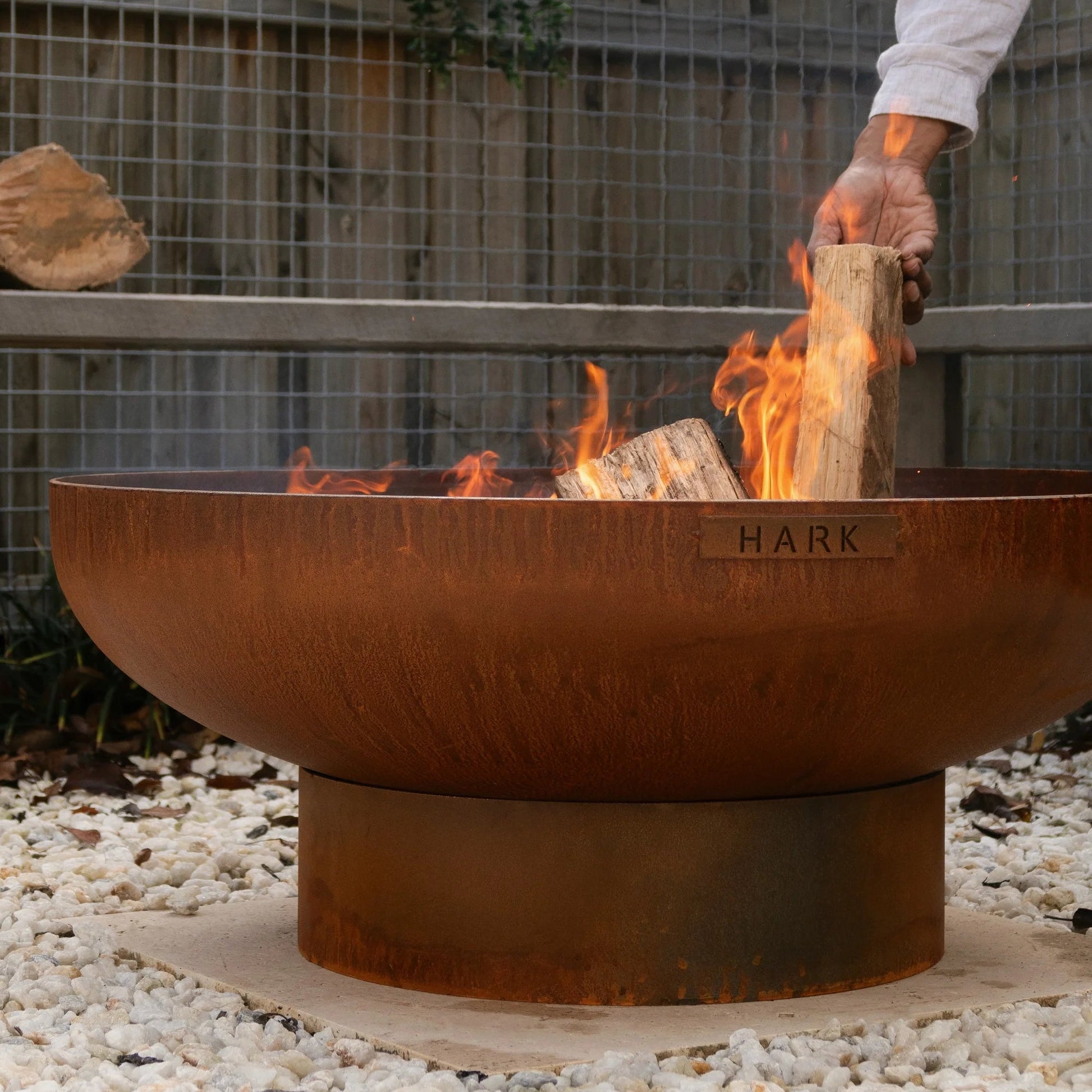 Person lighting wood in a rust-colored fire pit with a garden background
