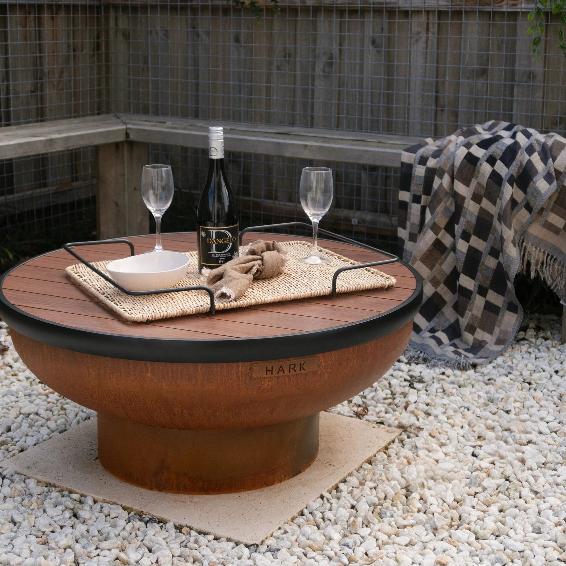 Outdoor setting with a fire pit table, wine glasses, and a bottle on a gravel surface.
