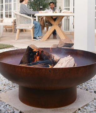 Rust-colored fire pit with flames on a pebble-covered patio, people sitting at a table in the background.