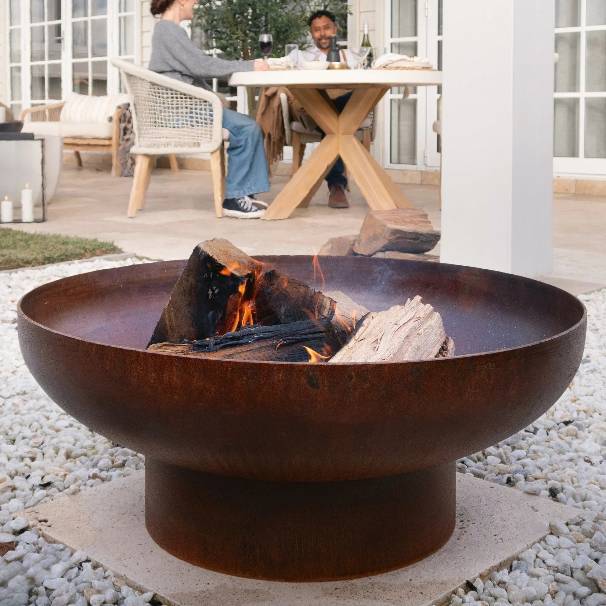 Rust-colored fire pit with flames on a pebble-covered patio, people sitting at a table in the background.
