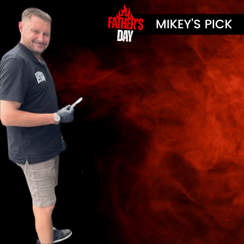A man wearing a dark navy polo shirt and gray shorts stands against a black background with a red smoke graphic, holding a lighter. Text at the top reads 'FATHER'S DAY MIKEY'S PICK' with a 'MIKEY'S PICK' logo.