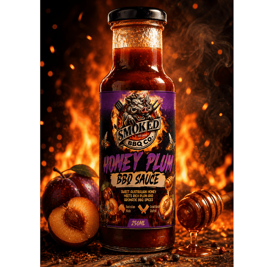 A bottle of Smoked BBQ Co Honey Plum BBQ Sauce stands on a rustic wooden surface with plums, a honey dipper, and glowing embers in the background. The bottle has a black lid and a purple label with fiery graphics.