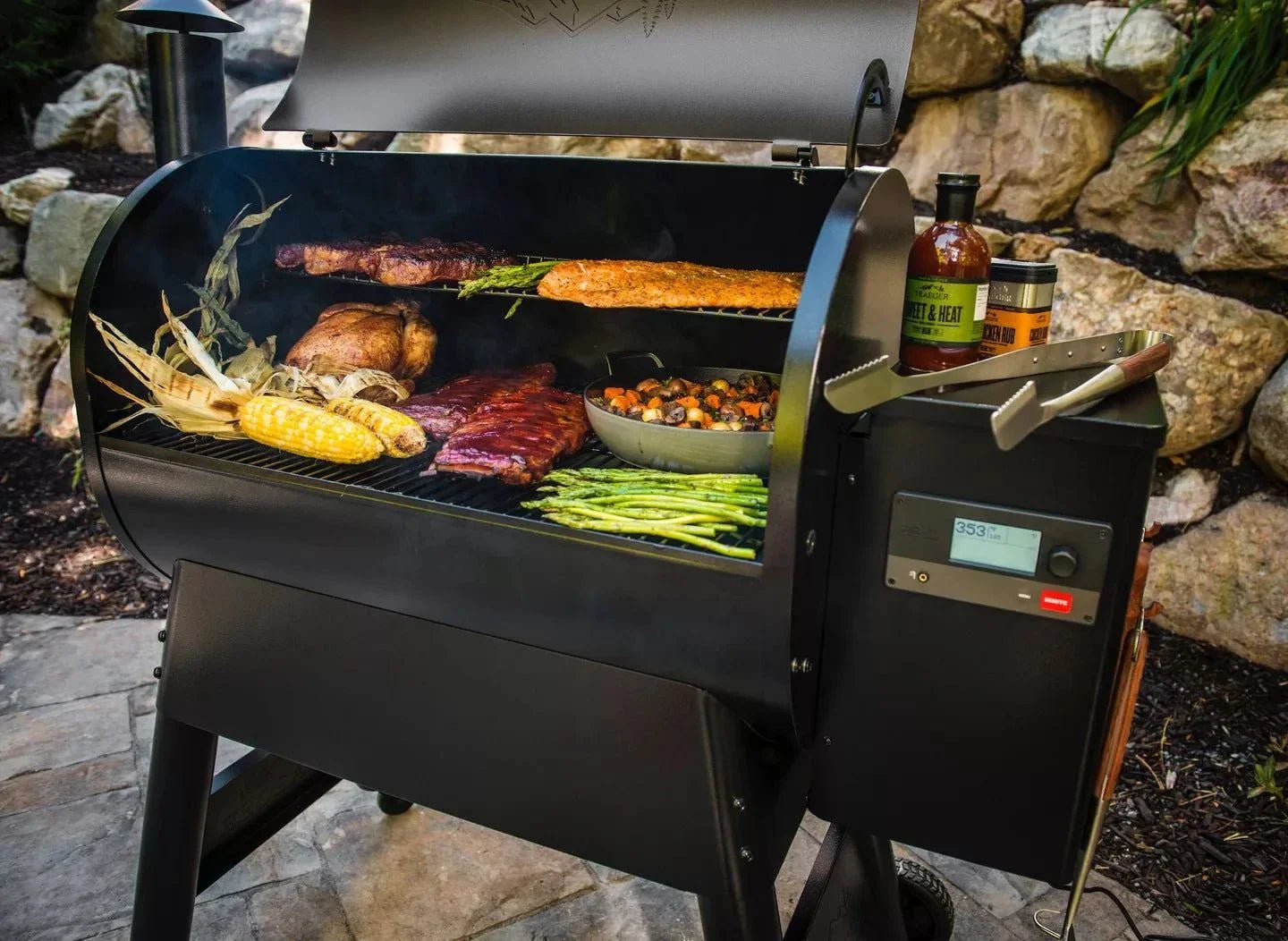 Black barbecue grill with food being cooked outdoors, surrounded by stones and plants.