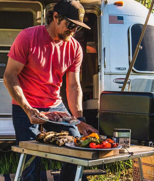Man grilling outdoors next to a trailer with food on a table