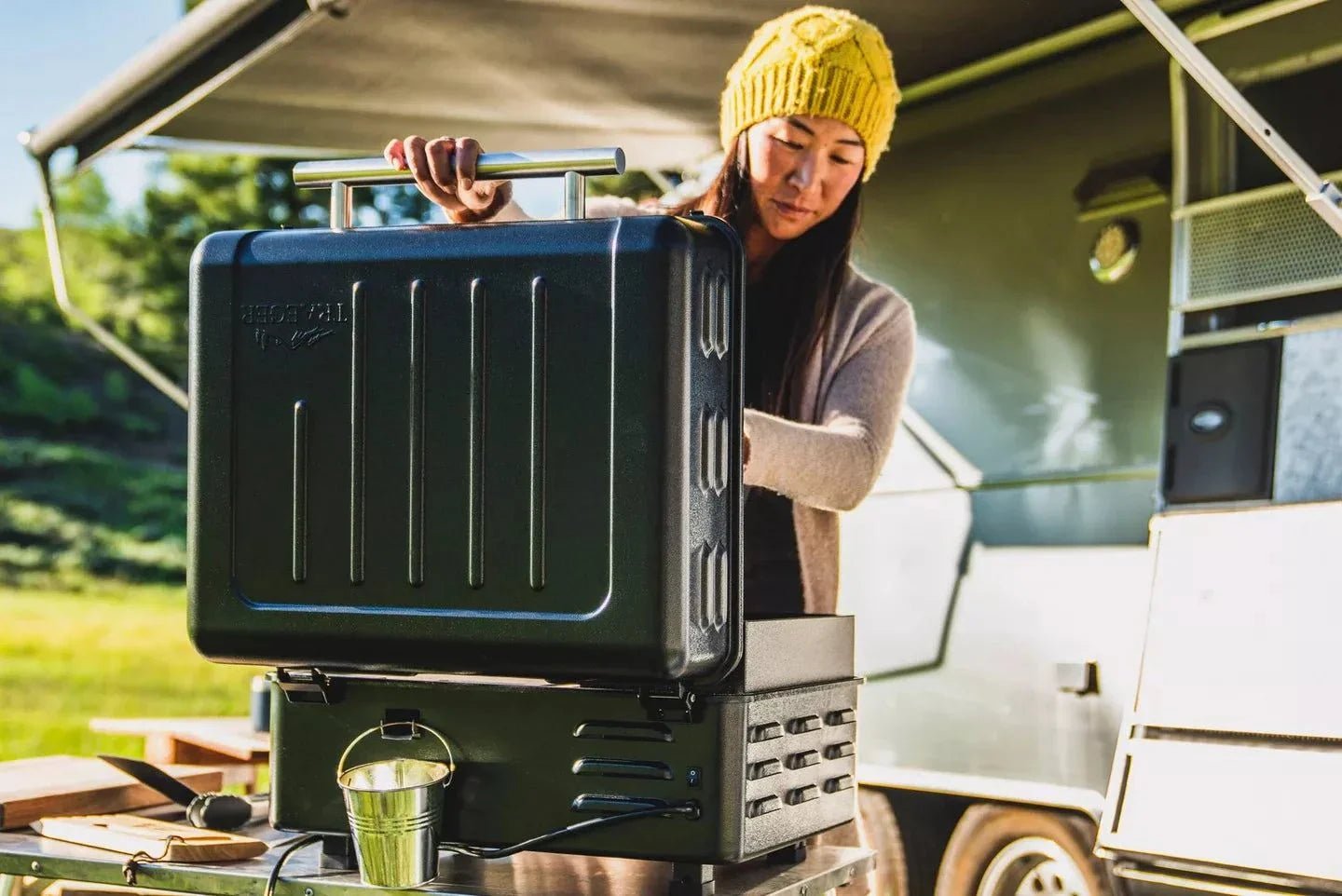 Person opening a black camping stove in front of a camper van