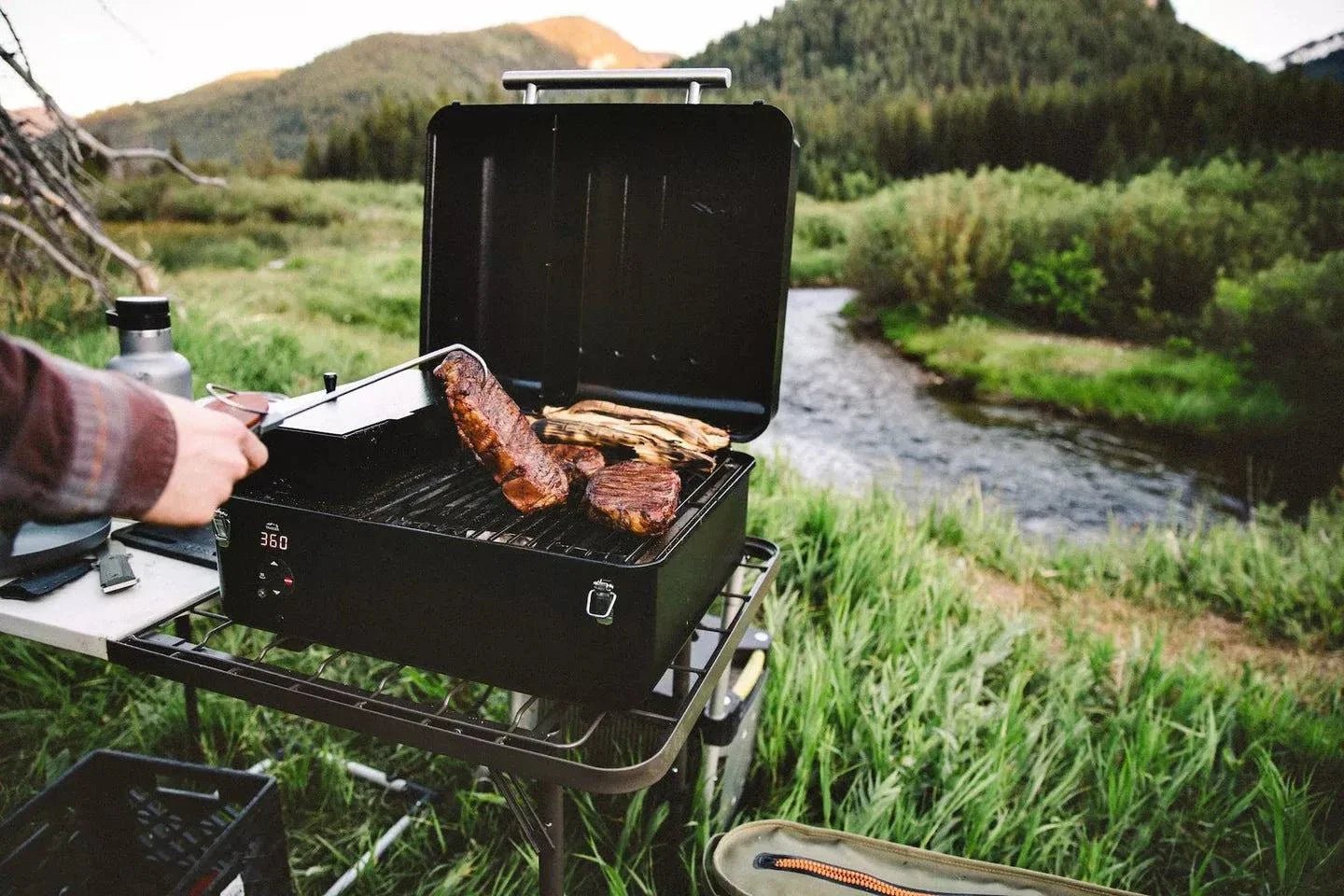Person grilling outdoors by a river with mountains in the background
