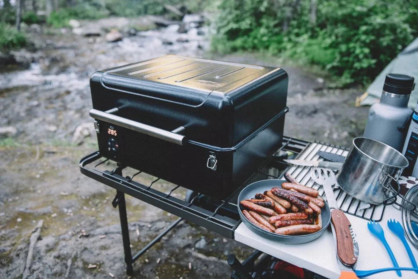 Portable grill with sausages on a camping setup by a stream